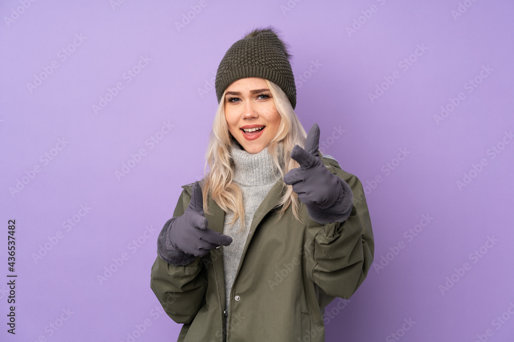 Teenager blonde girl with winter hat over isolated purple background pointing to the front and smiling Teenager blonde girl with winter hat over isolated purple background pointing to the front and smiling
