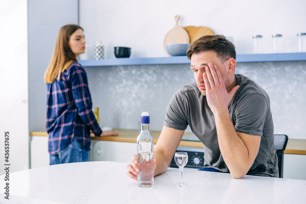 disgruntled housewife standing behind and watching her alcoholic husband. A couple of a man and a woman have conflicts in the home kitchen due to male alcoholism. Relationship and marriage problems