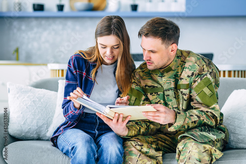 Canvastavla A young Caucasian civilian officer in camouflage uniform sits on a sofa in the living room with a girl showing a photo album, feeling peacefully happily reunited together