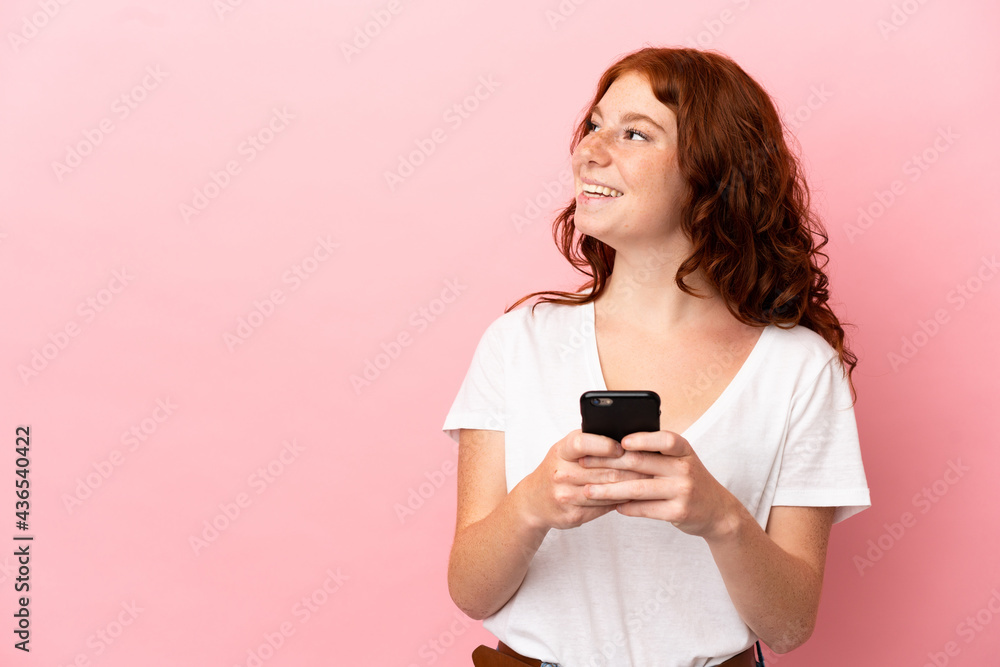 Teenager reddish woman isolated on pink background using mobile phone and looking up Teenager reddish woman isolated on pink background using mobile phone and looking up
