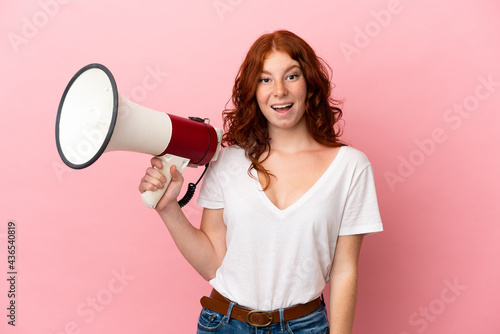 Teenager reddish woman isolated on pink background holding a megaphone and with surprise expression