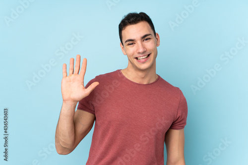 Young caucasian handsome man isolated on blue background saluting with hand with happy expression