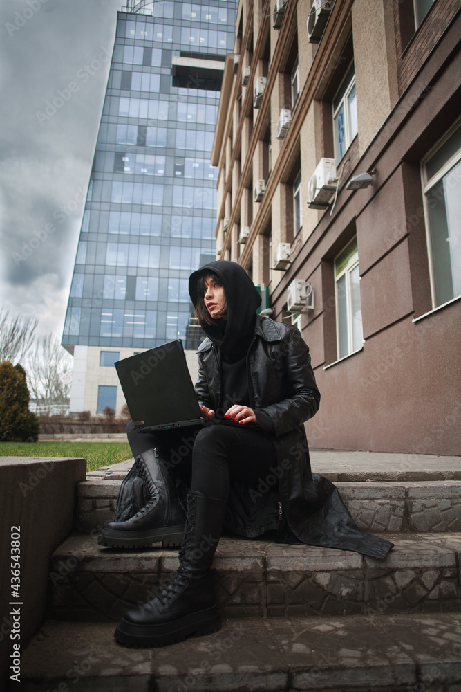 Girl in black leather coat and hood with laptop on city street, concept of woman hacker and agent working remotely at computer