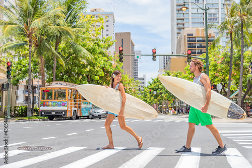 Honolulu Hawaii surfers couple tourists people walking crossing city street carrying surfboards going to the beach surfing. Surf living lifestyle. Surfer woman and man friends in Waikiki, Oahu, USA.