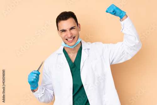 Young dentist man holding tools isolated on beige background celebrating a victory