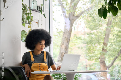 Young happy African American college student hipster girl with afro hair sitting at table in cafe indoor alone using laptop watching learning virtual online digital webinar.