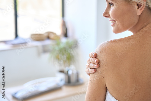 Fototapeta Back view of nude beautiful middle aged 50s woman applying exfoliating peeling sugar scrub after shower