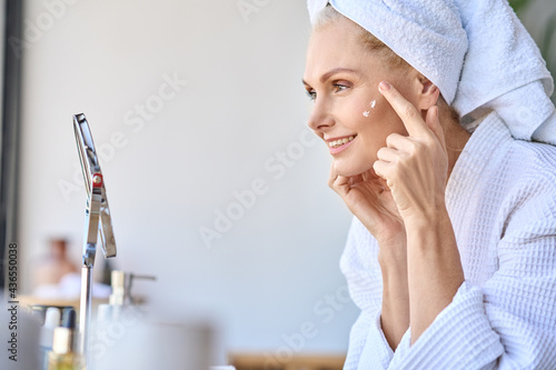 Gorgeous mid age older adult 50 years old blonde woman wears bathrobe in bathroom applying nourishing antiage face skin care cream treatment, looking at mirror doing daily morning beauty routine. photo