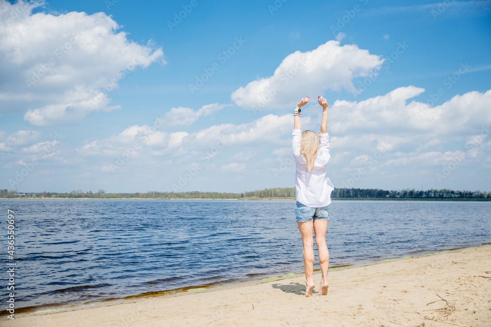 Young attractive blonde woman standing by lake with arms raised.