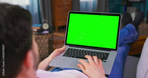 Man at Home Lying on a Couch Working on Green Mock-up Screen Laptop Computer. Guy Using Laptop Device, Browsing Internet, Watching Content, Videos. Over the Shoulder Camera Shot.