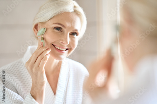 Senior mid aged lady of 50s years old massaging face skin using natural stone trendy jade quartz rolling device for detox of perfect skin. Spa traditional technique skincare ads.
