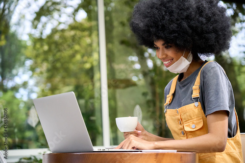 Smiling happy teenage African American hipster girl student sitting outdoor at cafe table using laptop watching online virtual webinar, working remote wearing face mask for covid 19 protection.