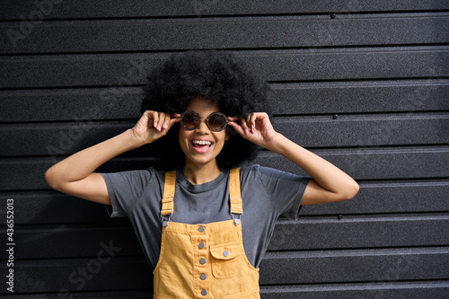 Portrait of young black afro American gen z laughing joyful model in her 20s. Funny mixed race teen lady wearing sun glasses standing on black wall background.