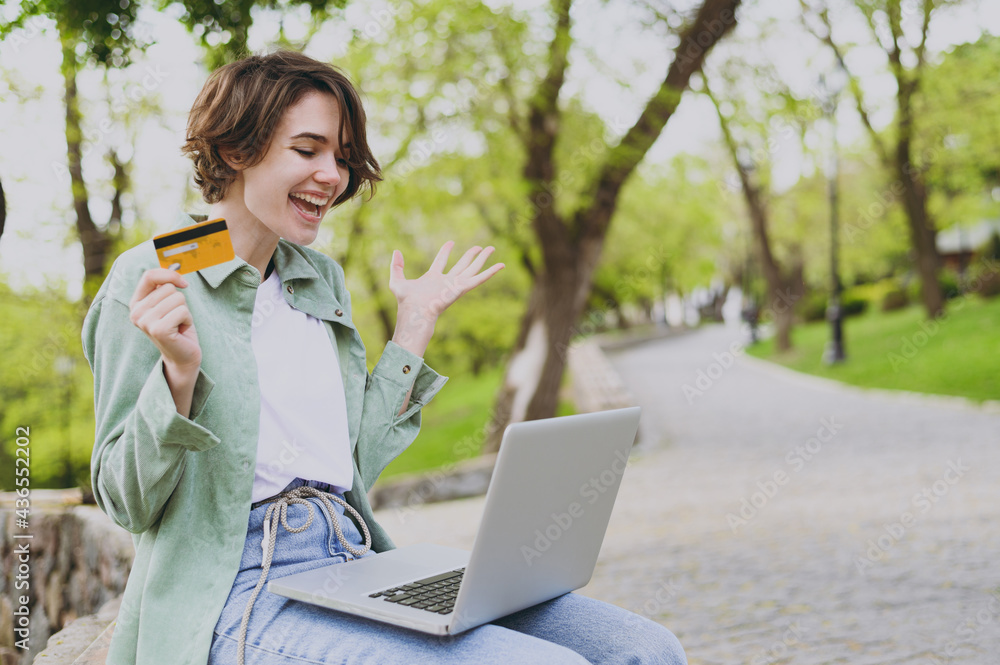 Young surprise woman in green jacket sit on bench in park outdoors rest use laptop pc computer hold credit bank card do online shopping order booking tour spread hands People urban lifestyle concept Young surprise woman in green jacket sit on bench in park outdoors rest use laptop pc computer hold credit bank card do online shopping order booking tour spread hands People urban lifestyle concept