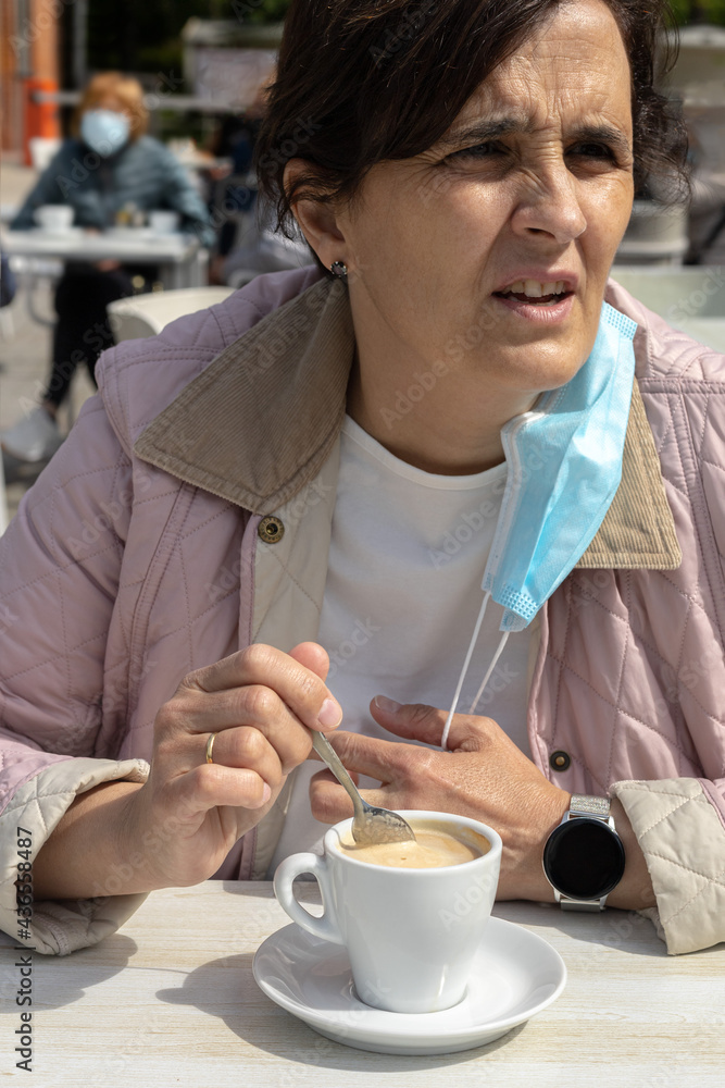 Young adult woman drinking a latte on the terrace of a coffee shop and pulling her coronavirus protection mask away with a finger Young adult woman drinking a latte on the terrace of a coffee shop and pulling her coronavirus protection mask away with a finger