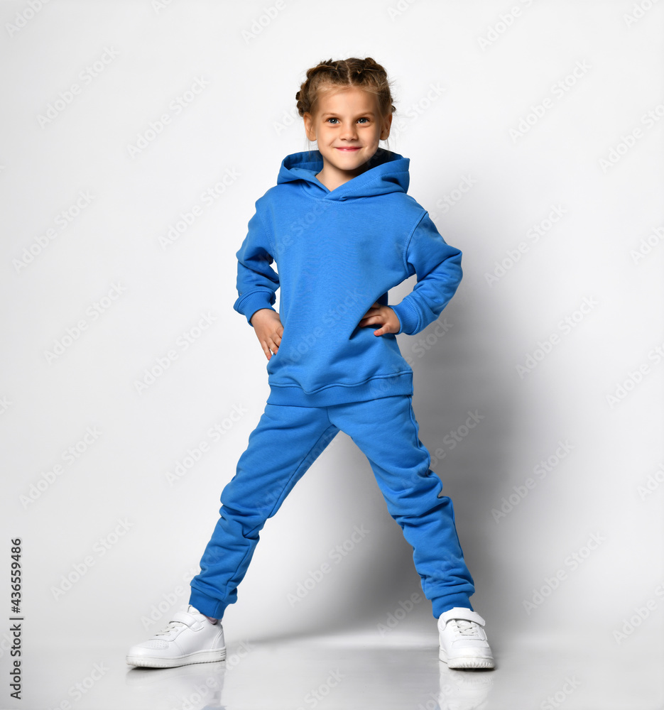 little girl in a trendy blue tracksuit having fun on a white background. A child with an emotional expression is standing with his hands on his waist. Promotion of children's sports fashion. little girl in a trendy blue tracksuit having fun on a white background. A child with an emotional expression is standing with his hands on his waist. Promotion of children's sports fashion.