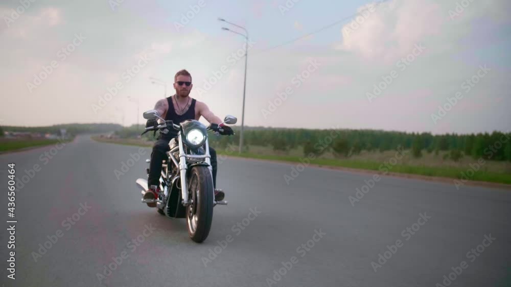 Stylish biker with tattoos rides a motorcycle on a country road at sunset Stylish biker with tattoos rides a motorcycle on a country road at sunset
