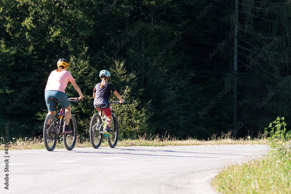Woman and girl child riding mountain bikes on a country road in nature on a bright sunny summer day.