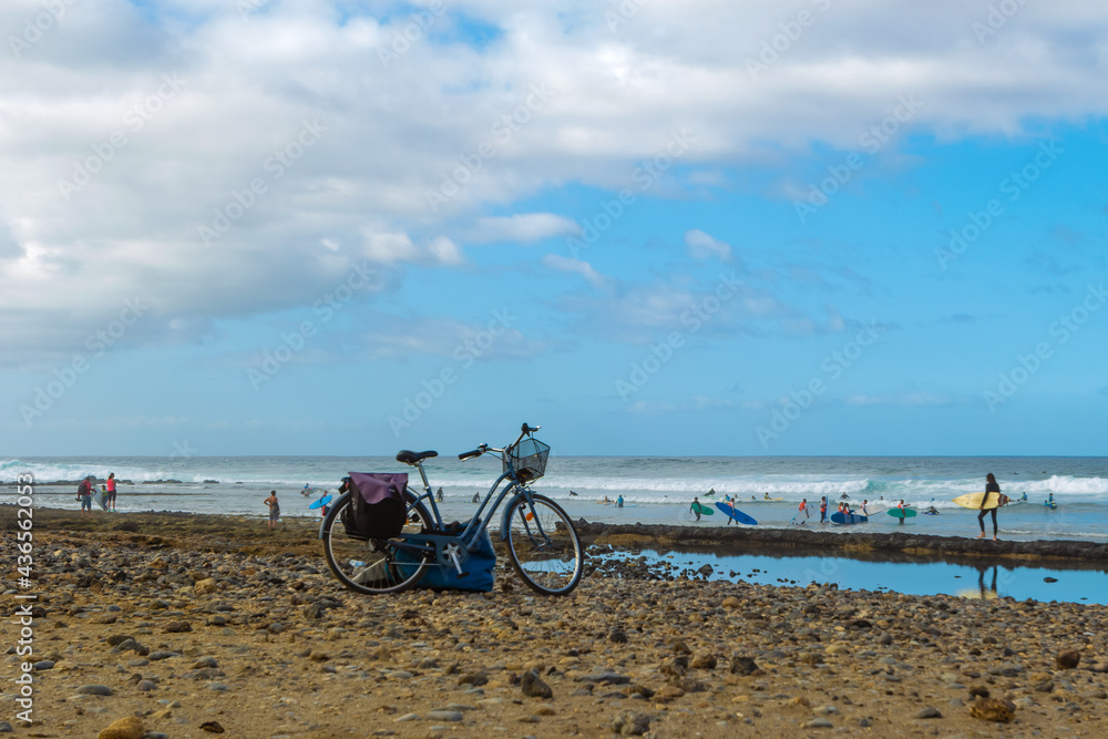 The bike stands on the beach and surfers on the ocean. Santa Cruz de Tenerife, Spain’s Canary Island