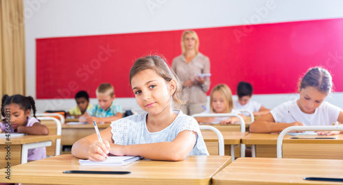 Focused girl sitting at desk writing test in classroom full of pupils during lesson