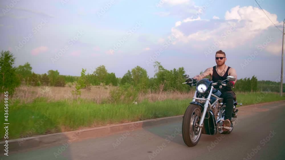 Stylish biker with tattoos rides a motorcycle on a country road at sunset Stylish biker with tattoos rides a motorcycle on a country road at sunset