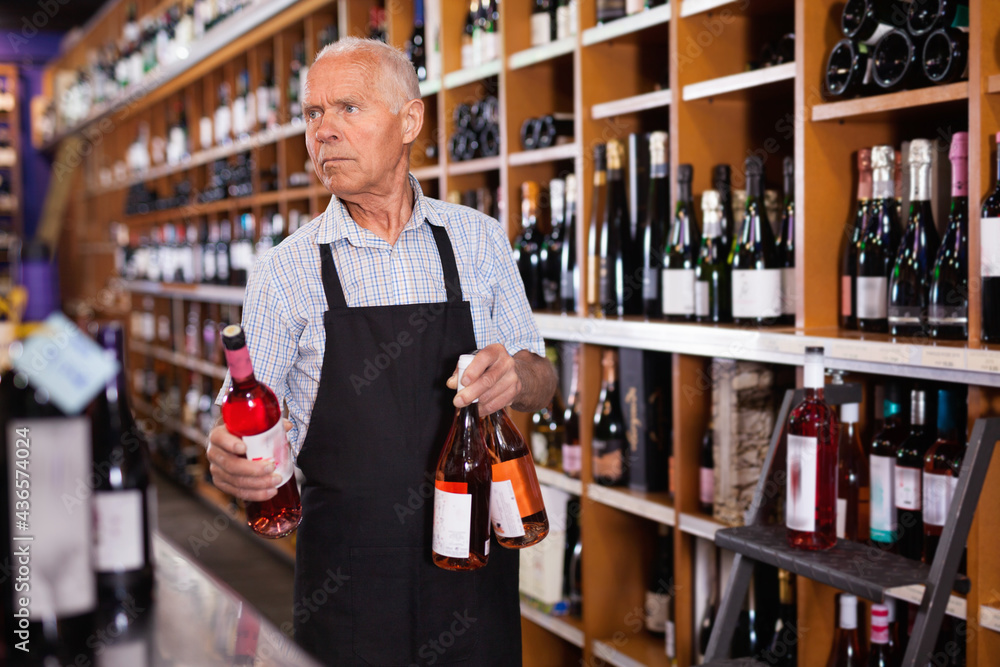 Confident elderly cheerful salesman of wine house arranging wine bottles on shelves rack
