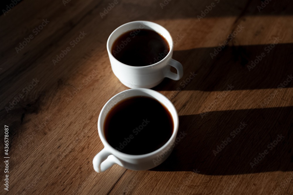 Close-up of two white ceramic cups of coffee with shadows on wooden table. Close-up of two white ceramic cups of coffee with shadows on wooden table.