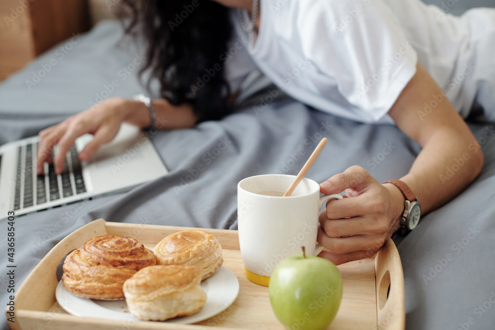 Close-up of unrecognizable man lying on bed and using laptop, he having breakfast in bed while working at home Close-up of unrecognizable man lying on bed and using laptop, he having breakfast in bed while working at home