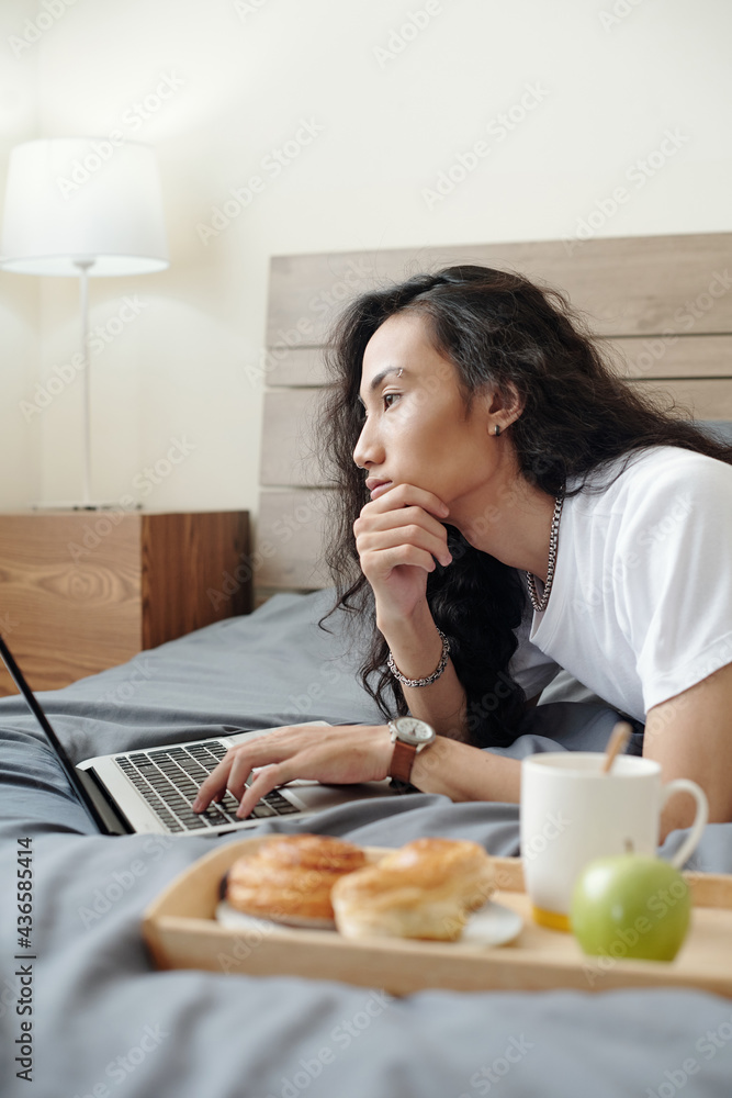 Pensive young Vietnamese online manager with long hair lying on bed with breakfast on tray and using laptop while thinking of project Pensive young Vietnamese online manager with long hair lying on bed with breakfast on tray and using laptop while thinking of project