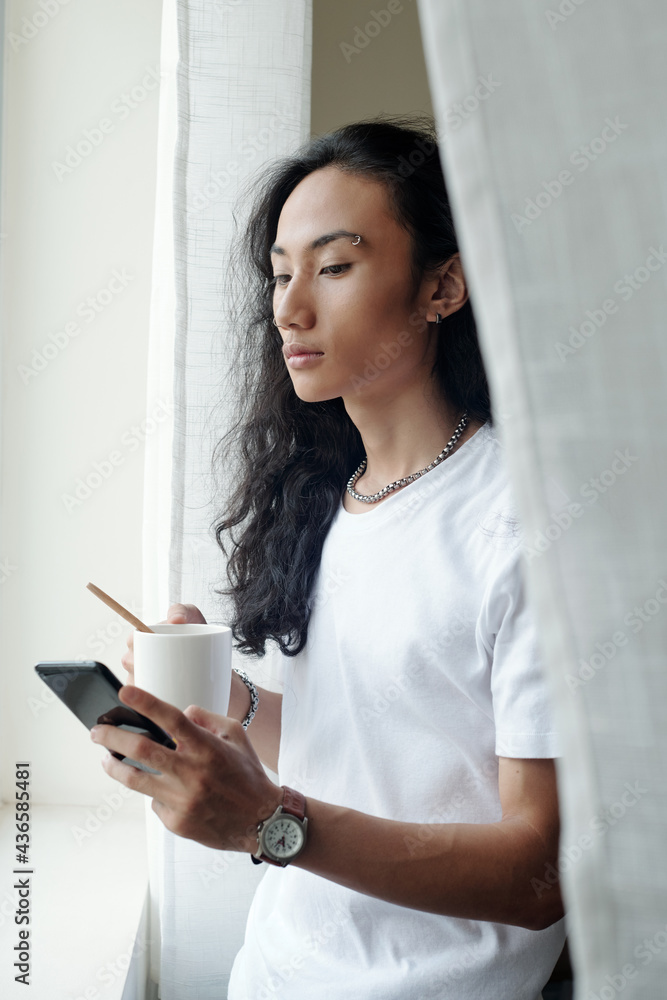 Serious young Vietnamese man with wavy hair standing at window and drinking coffee while scrolling through social media Serious young Vietnamese man with wavy hair standing at window and drinking coffee while scrolling through social media