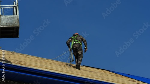 Wallpaper Mural Builder with safety harness and helmet walks holding drill along renewing roof against blue sky on sunny day backside view Torontodigital.ca