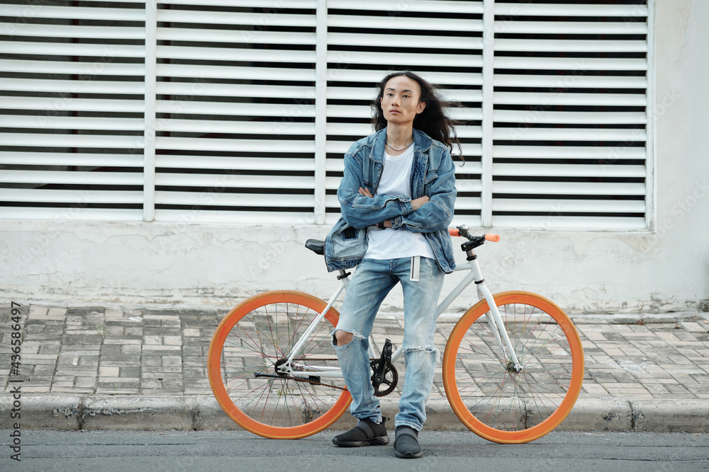 Serious confident young Asian bicyclist in denim wear standing with crossed arms against bicycle with orange wheels outdoors Serious confident young Asian bicyclist in denim wear standing with crossed arms against bicycle with orange wheels outdoors