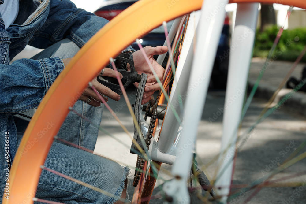 Close-up of unrecognizable man in denim wear crouching on asphalt and installing bike pedal outdoors