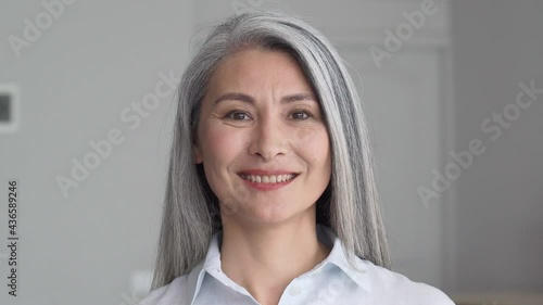 Happy smiling stylish confident 50 years old Asian female professional standing looking at camera at gray background. Portrait of sophisticated grey hair woman advertising products and services.