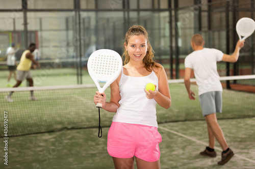 Portrait of smiling sporty girl with paddle tennis racquet and ball in her hands ready to play match standing on indoor court on blurred background of opponents © JackF