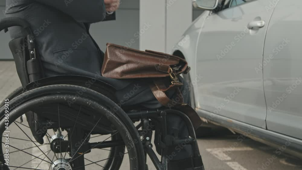Mid-section tracking of unrecognizable disabled businessman in wheelchair riding towards his car, then unlocking doors and putting his briefcase into driver seat Mid-section tracking of unrecognizable disabled businessman in wheelchair riding towards his car, then unlocking doors and putting his briefcase into driver seat