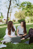 Two female students are sitting in the park on the grass with books and laptops, studying and preparing for exams. Distance education. Soft selective focus.