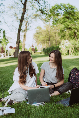 Two female students are sitting in the park on the grass with books and laptops, studying and preparing for exams. Distance education. Soft selective focus. © Tasha Sinchuk