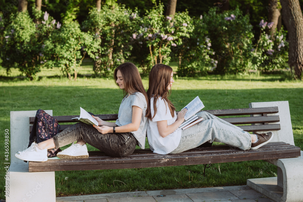 Two student girls are looking at an open book on a bench in the park. Distance education, preparation for exams. Soft selective focus. Two student girls are looking at an open book on a bench in the park. Distance education, preparation for exams. Soft selective focus.