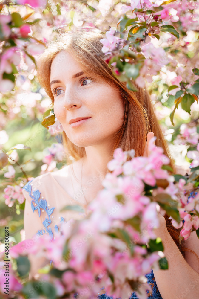 Portrait of a red-haired spring girl. Beautiful young woman in pink flowers enjoying the apple orchard. Allergy free concept.