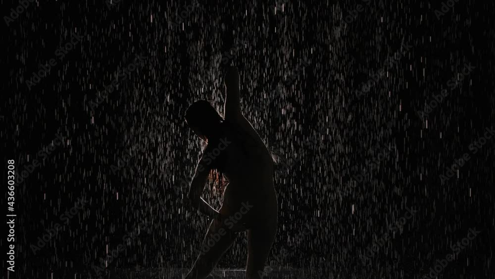 Modern ballet, contemp, contemporary performed by woman in rain against backdrop of bright spotlight. Silhouette of dancer moving in slow motion among shiny drops of rain. Modern ballet, contemp, contemporary performed by woman in rain against backdrop of bright spotlight. Silhouette of dancer moving in slow motion among shiny drops of rain.