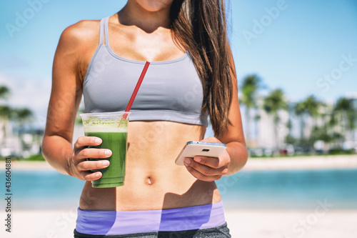 Healthy green smoothie juice drink online order on phone app fit athlete girl drinking detox cleanse morning breakfast on beach. Sport woman outside in summer sun with diet food.