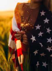 Young woman holding bengal fire with American flag at sunset. America celebrate 4th of July. Independence Day. 	
