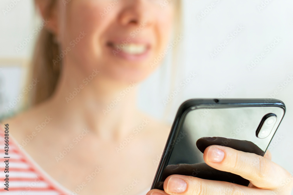 Smiling young woman using phone at home. Focus on the smartphone. A blond hair girl holds a cell phone in her hands, checking online news and social networks, orders delivery. Technology concept