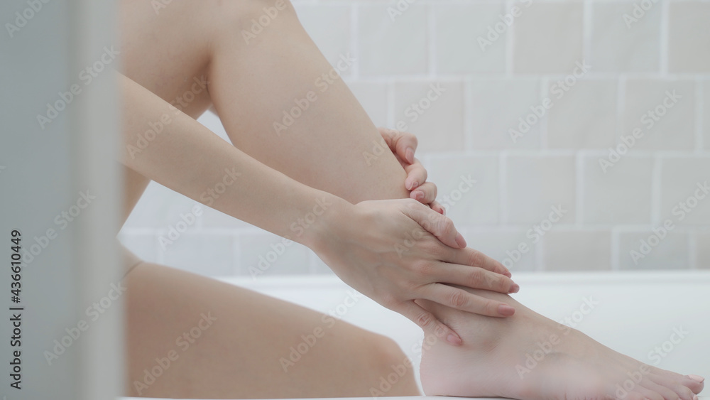 Fototapeta premium Asian woman short hair, applying cream lotion on hands, leg, and arm in white bathroom near bathtub at home studio decor some green plant. white cream rubbing to treatment skin condition. scrubbing