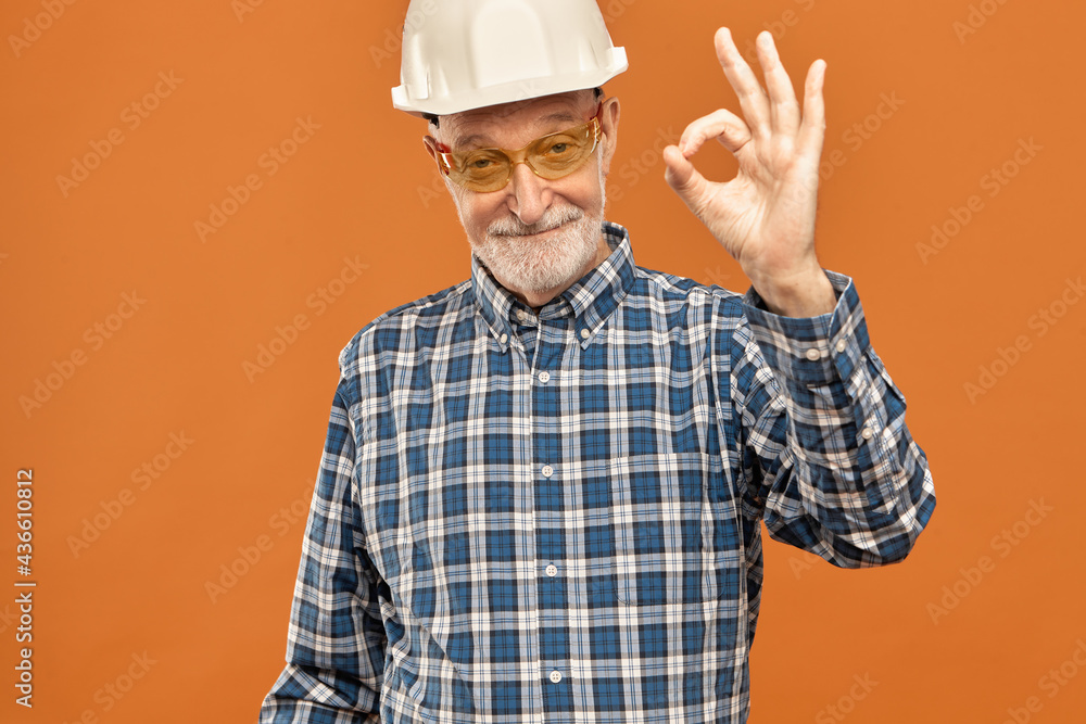 Naklejka premium Skilled positive mature bearded male supervisor in white hard hat and safety glasses approving plan of construction project showing okay gesture, saying everything is perfect, smiling joyfully