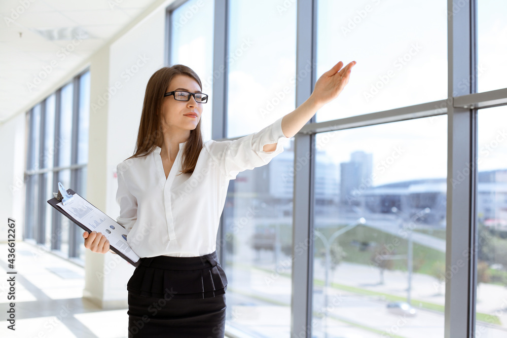Beautiful female specialist with clipboard standing in modern office and smiling charmingly. Working on design, data analysis, plan strategy. Business people concept