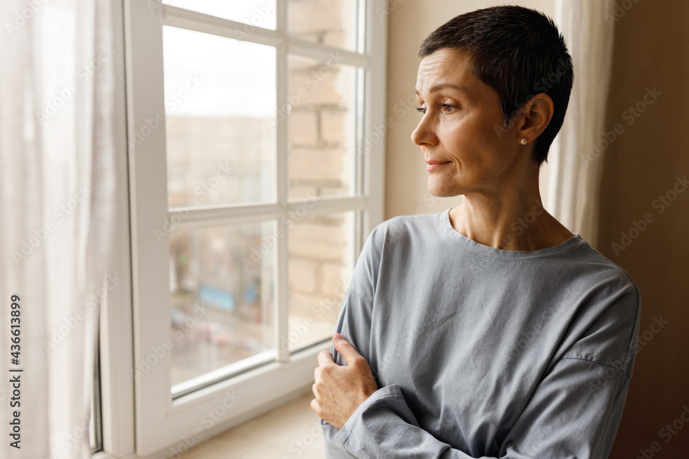 Portrait of sad middle aged female with short hairstyle posing by window looking outside with bored pensive facial expression, crossing arms on her chest. People, home and domestic life concept Portrait of sad middle aged female with short hairstyle posing by window looking outside with bored pensive facial expression, crossing arms on her chest. People, home and domestic life concept