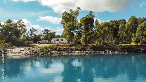 Landsscape view of a large modern caravan and four wheel drive vehicle free camping alongside a beautiful blue lake fringed with gumtrees.