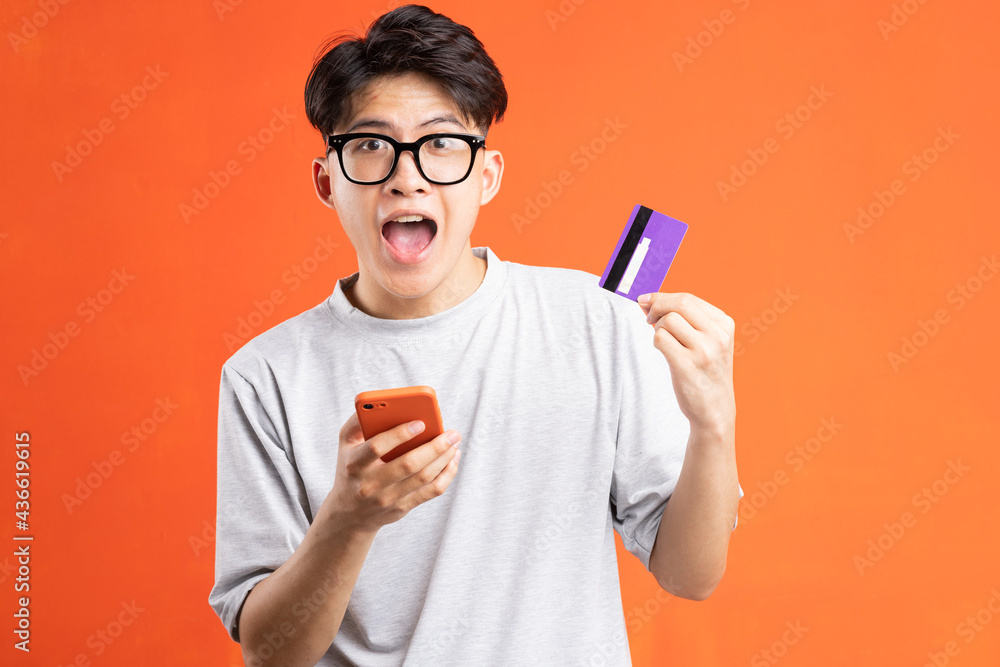 Portrait of young asian man holding smart phone and ATM card, isolated on orange background Portrait of young asian man holding smart phone and ATM card, isolated on orange background